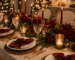 Mesa de Natal sofisticada com toalha neutra, louças brancas, guardanapos vinho presos por laços de veludo vermelho, flores marsala, folhagens verdes, pinhas, talheres dourados e iluminação quente.