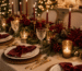 Mesa de Natal sofisticada com toalha neutra, louças brancas, guardanapos vinho presos por laços de veludo vermelho, flores marsala, folhagens verdes, pinhas, talheres dourados e iluminação quente.