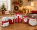 Composição de peças natalinas de cerâmica esmaltada em tons de branco, vermelho e dourado, incluindo Papai Noel, pequenas casas e árvores sobre um aparador elegante.