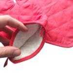 Pessoa utilizando a luva térmica natalina para segurar uma assadeira, demonstrando o formato da peça e sua aplicação durante o preparo de alimentos.